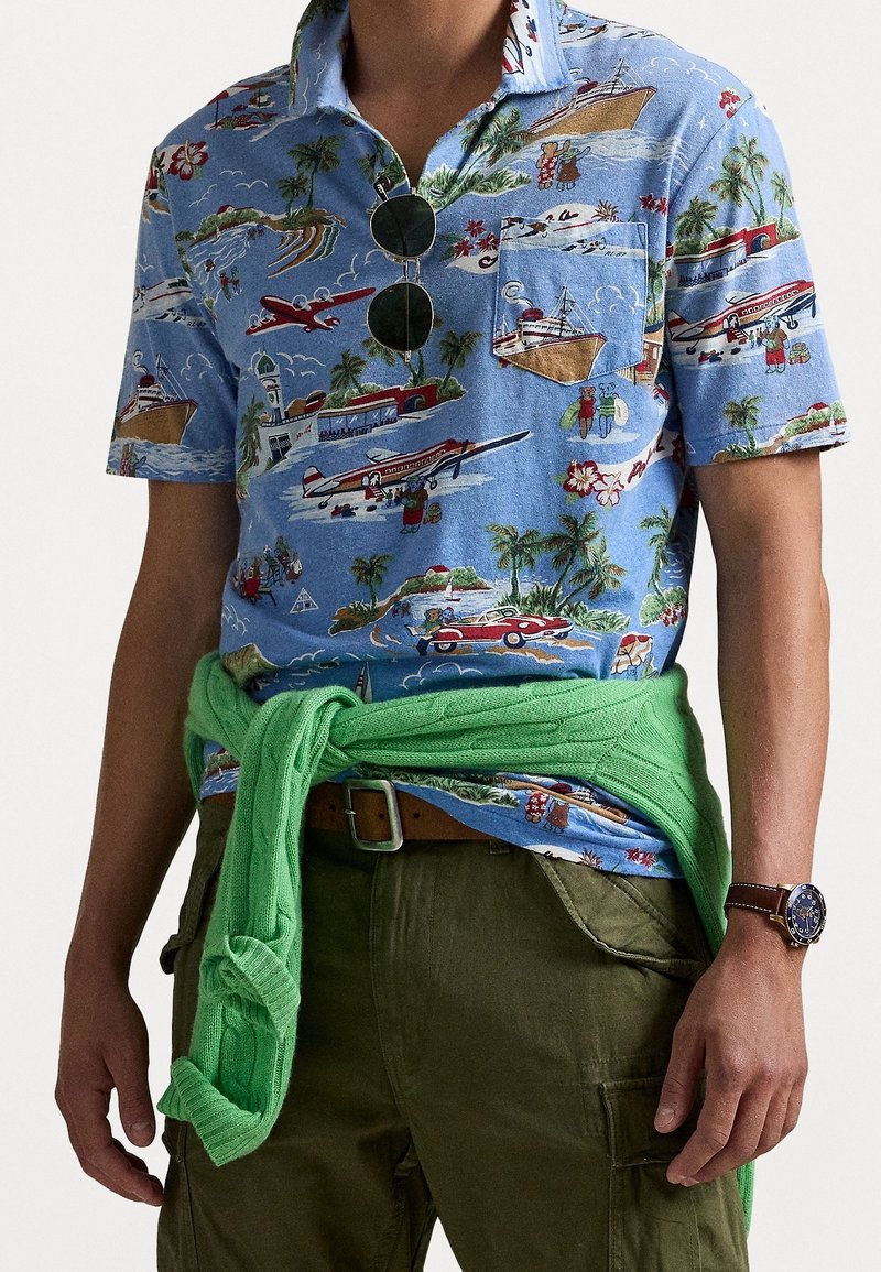 Homme portant une chemise bleue à manches courtes avec un imprimé tropical de voyage, un pull vert noué autour de la taille, un pantalon olive, des lunettes de soleil rondes et une montre-bracelet.
