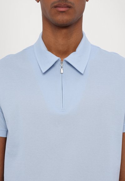 Ανοιχτό μπλε πόλο μπλούζα με υφασμάτινη υφή, κοντά μανίκια και μισό φερμουάρ στο γιακά. Απλός σχεδιασμός χωρίς σχέδια ή διακοσμήσεις.