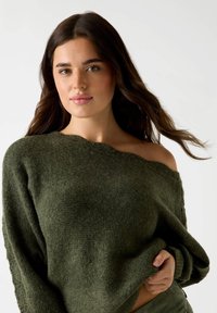 Kobieta z długimi ciemnymi włosami, ubrana w ciemnozielony sweter odsłaniający ramiona, trzymająca brzeg swetra jedną ręką.