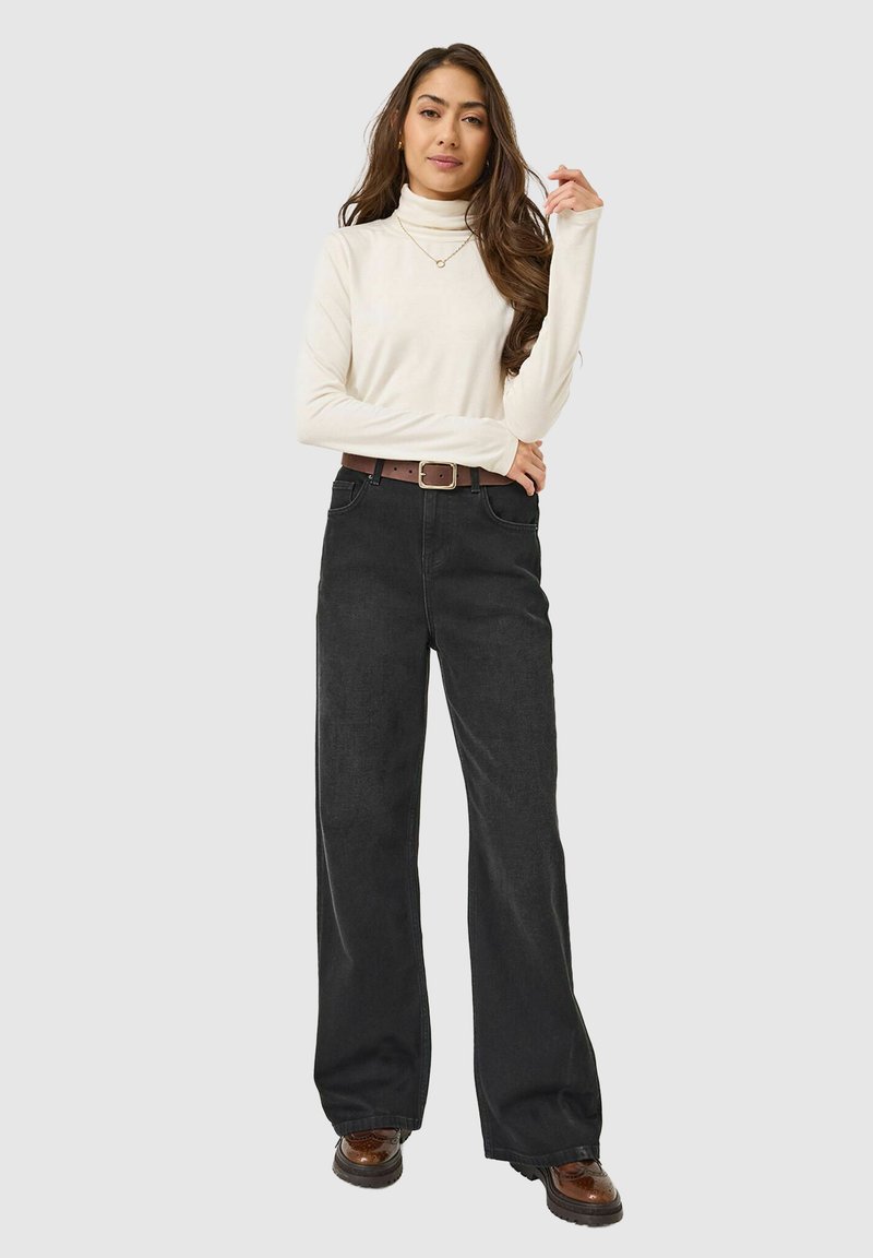 Femme avec de longs cheveux bruns portant un col roulé blanc, un jean noir à jambes larges avec une ceinture marron et des chaussures marron, se tenant les bras croisés.