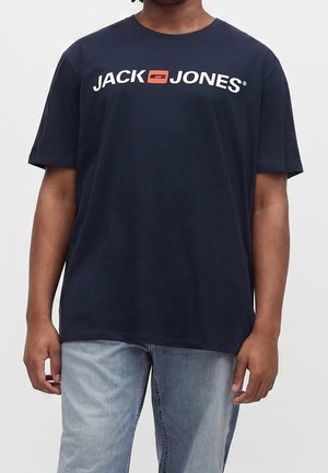Person iført en mørkeblå Jack & Jones t-shirt og lyseblå jeans, stående mod en ensfarvet lys baggrund.