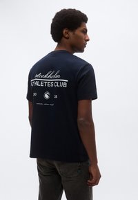 Tengerészkék pamut póló, környakkivágással, rövid ujjakkal, a háton a "Stockholm Athletes Club" feliratot és egy logót ábrázoló nyomott mintával.