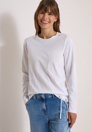 Femme aux cheveux bruns portant une chemise blanche à manches longues et un jean bleu, souriante avec une main dans la poche sur un fond uni.