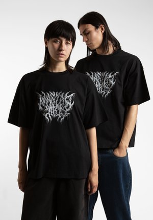 Schwarze, übergroße T-Shirts mit auffälligem, tropfendem weißen Grafikdesign auf der Vorderseite. Models tragen lässig sitzende Denim und dunkle Hosen.