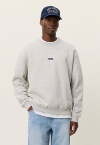 Άνδρας με ανοιχτό γκρι φούτερ με μικρό λογότυπο, ανοιχτό μπλε τζιν και σκούφο ναυτικού μπλε, στέκεται μπροστά σε απλό φόντο.