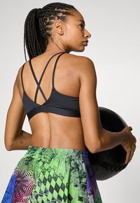 Soutien-gorge de sport noir avec un design à dos croisé, associé à un short coloré et à motifs avec des tons verts, violets et noirs. Tenant un ballon de médecine.