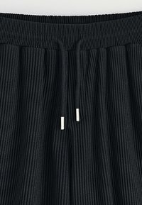 Pantalons en tissu noir côtelé avec une taille élastique, dotés d'un cordon de serrage avec des embouts métalliques pour un ajustement réglable. Design texturé sur l'ensemble.