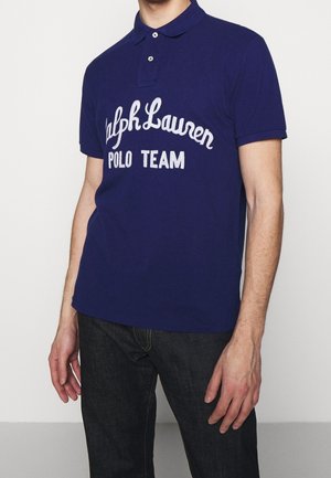 Homme portant un polo bleu foncé avec l'inscription "Ralph Lauren POLO TEAM" brodée en blanc, associé à un jean foncé, debout devant un fond blanc.