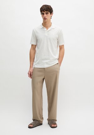 Junger Mann, der ein weißes Polo mit kurzen Ärmeln, beige weite Hosen und graue offene Hausschuhe vor einem einfarbigen Hintergrund trägt.