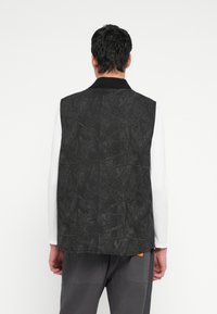 Μαύρο γιλέκο με υφασμάτινο, αφηρημένο μοτίβο· διαθέτει γιακά και στιβαρό στρώμα, φορεμένο πάνω από ένα λευκό πουκάμισο με μακριά μανίκια.