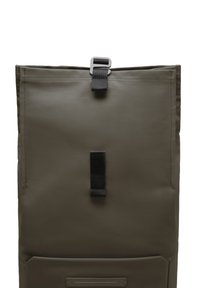 Sac à dos à rabat de couleur vert olive fabriqué à partir d'un matériau texturé. Il dispose d'une fermeture à boucle noire et d'une poche avant avec une plaque mince et rectangulaire.