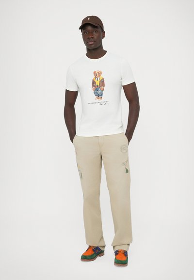 T-shirt branca de algodão com um gráfico colorido de um urso, calças bege com vários bordados decorativos e sapatos multicoloridos com acabamento texturizado.