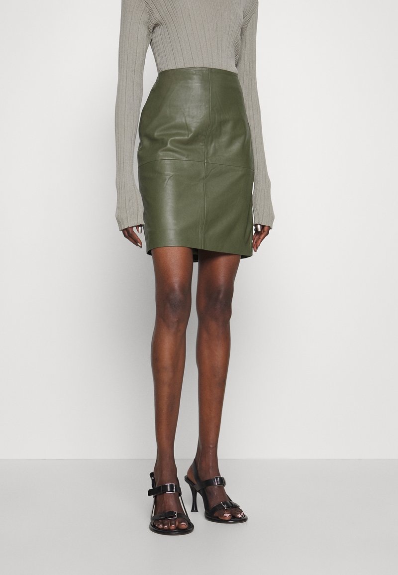 Kvinde iført en olivengrøn lædernederdel, beige ribbet langærmet top og sorte åbne hæle, der står på et ensfarvet gulv.