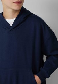 Felpa in cotone blu navy con scollo a V, maniche lunghe e tasca a marsupio frontale con polsini a coste. Texture liscia e stile minimalista.
