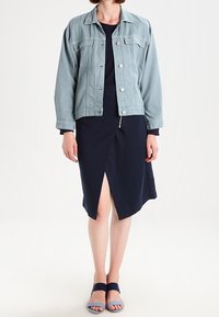 Chaqueta de mezclilla azul clara, vestido navy con abertura frontal y sandalias azul bicolor. La chaqueta tiene un collar clásico y botones.
