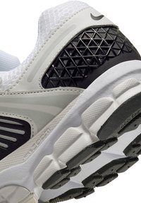 Nike športni copat z belim mrežastim zgornjim delom, črnim geometrijskim dodatkom na peti, teksturiranim podplatom in gumijastim podplatom z izrazitim vzorcem.