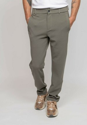 Homme portant un pantalon slim vert olive avec poches et des baskets beige, les mains dans les poches, debout devant un fond neutre.