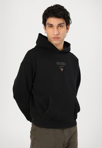 Čierna mikina s kapucňou vyrobená z bavlny, so prednou kapsou a kapucňou na šnúrku. Na hrudi vyšívané logo "GUESS" v tmavom zlate.