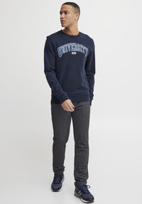 Marinblå sweatshirt med "UNIVERSITY" i ljusblå bokstäver, i kombination med mörkgrå slim-fit byxor och marinblå sneakers. Ser avslappnad och bekväm ut.