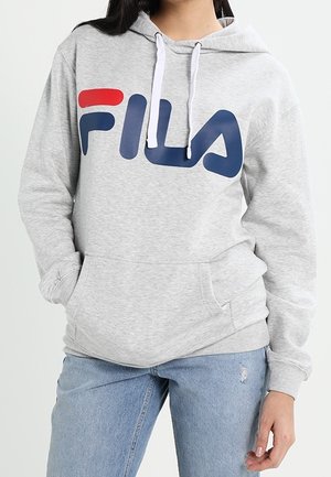 Personne portant un sweat à capuche FILA gris clair avec des cordons blancs et un jean bleu avec une petite déchirure près du genou, mains dans les poches.