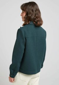 Veste en laine mélangée vert foncé, au design structuré avec un col, des poignets boutonnés et des détails sur le dos. Semble chaude et texturée.