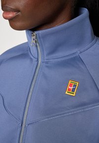 Chaqueta azul pálido con cremallera, con un cuello alto, tela texturizada y un pequeño logotipo bordado multicolor en el pecho.