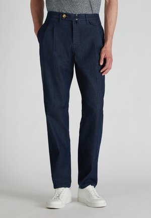 Marineblaue, plissierte Hose aus Denim mit glatter Textur. Verfügt über einen Knopfverschluss und Seitentaschen, kombiniert mit weißen Sneakers.