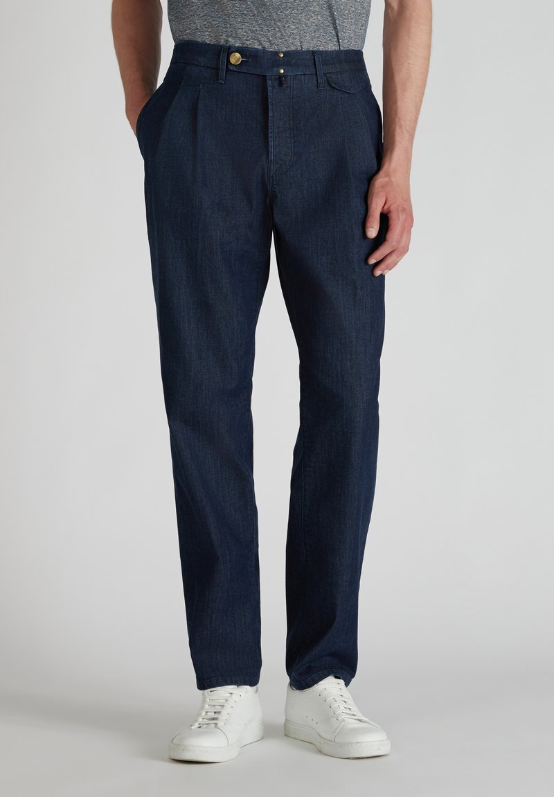 Navyblauwe geplooide broek van denim met een gladde textuur. Voorzien van een knoopsluiting en zijzakken, gedragen met witte sneakers.