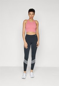 Jasnoróżowy prążkowany top crop zestawiony z ciemnymi legginsami z szarymi akcentami. Modelka nosi białe buty sportowe. Prosty, nowoczesny strój do ćwiczeń.