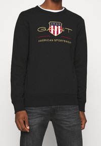 Sudadera negra con un logo bordado en forma de escudo, con rayas rojas y blancas, letras doradas y cuello redondo.