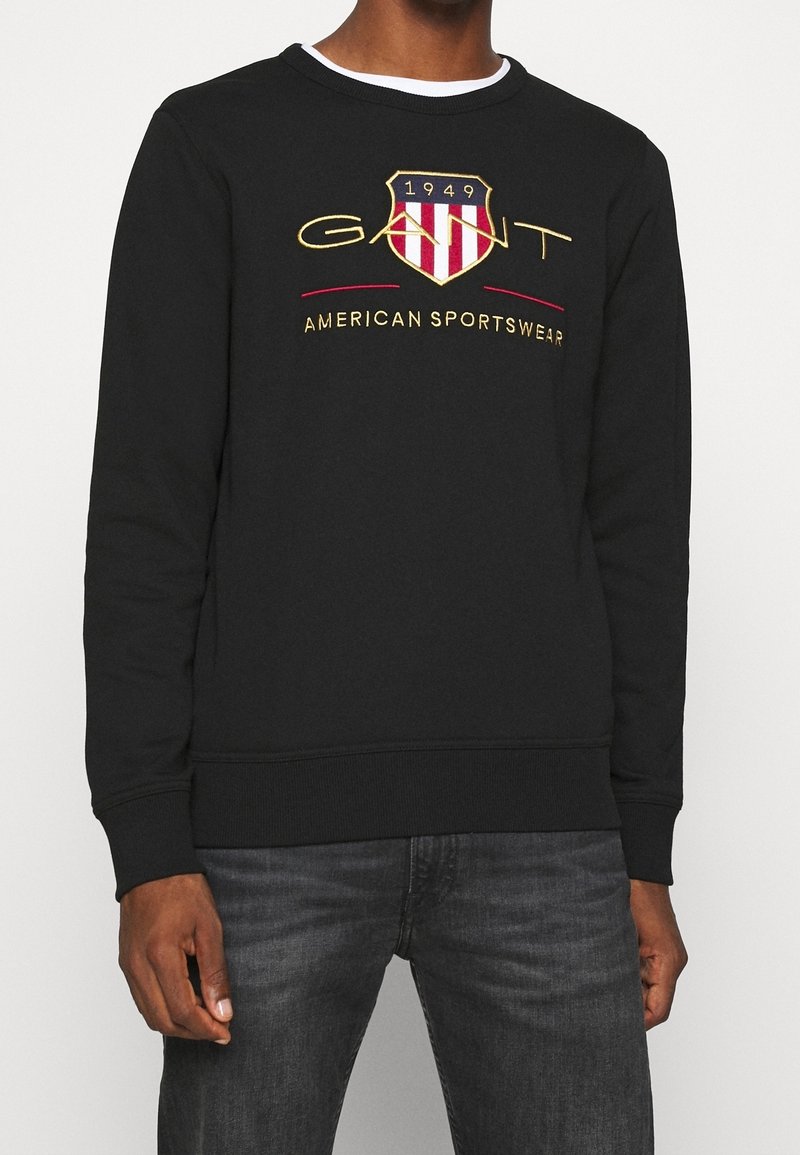 Sudadera negra con un logo bordado en forma de escudo, con rayas rojas y blancas, letras doradas y cuello redondo.
