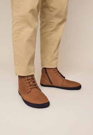 Person trägt beige Hose und braune Leder-Stiefeletten mit schwarzen Sohlen und seitlichen Reißverschlüssen, steht auf einer beigen Fläche.