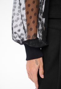 Czarna siateczkowa rękaw z okrągłymi wzorami, prążkowany czarny mankiet oraz tatuaż na dłoni opierającej się na ciemnej tkaninie. Przezroczysty i teksturowany wzór.