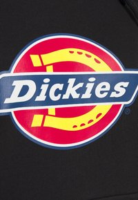 Logo de la marque Dickies avec texte blanc sur une bannière bleue au-dessus d'un cercle rouge comportant un design de fer à cheval jaune, sur un fond de tissu noir.
