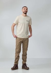 Beige tričko s logem, hnědé kalhoty a hnědé šněrovací boty. Kalhoty mají rovný střih; látka vypadá odolně a texturovaně.