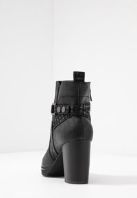 Botín negro con acabado en cuero texturizado, tacón prominente, correa decorativa con hebilla y parte trasera suave. Presenta detalles de costura.