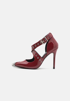 Chaussure à talon haut en cuir verni rouge avec bout pointu en métal, double brides croisées, boucle réglable et détails en œillets métalliques.