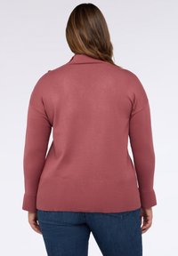 Rosa gestrickter Pullover mit hohem Kragen, gerippten Bündchen und Saum. Enthält Knopfakzente an der Schulter, kombiniert mit dunklen Jeans.