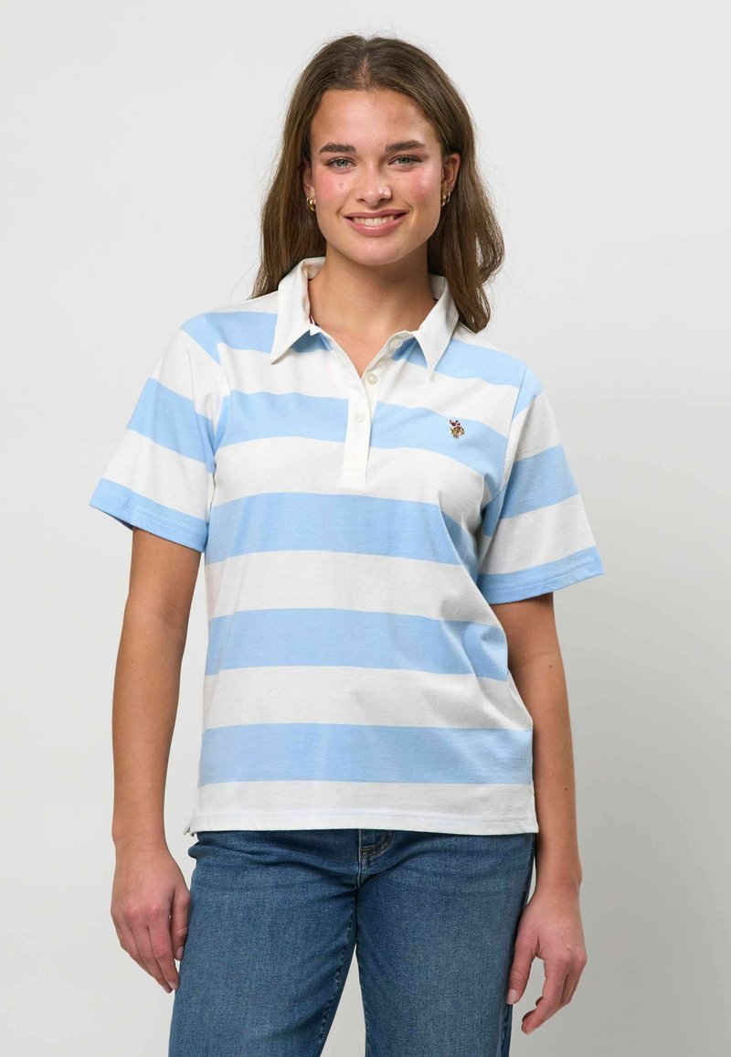 Femme aux cheveux bruns portant un polo à manches courtes rayé bleu et blanc et un jean bleu, debout devant un fond clair uni.