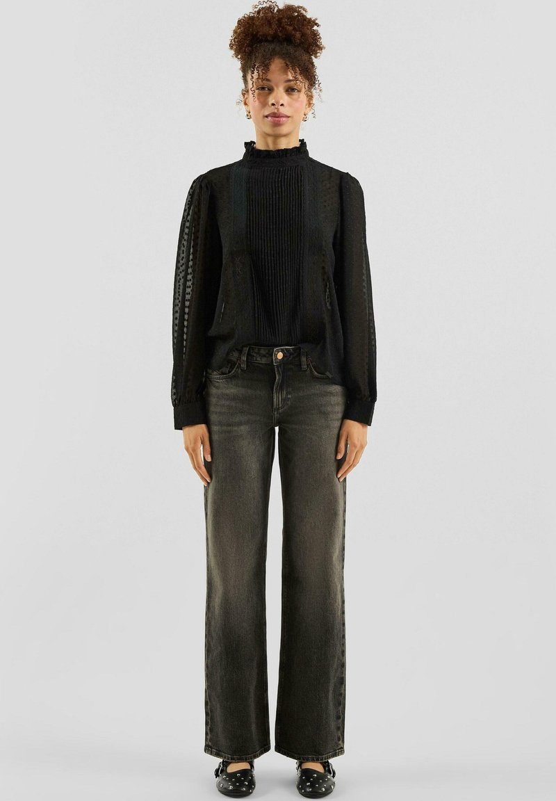 Zwarte blouse met gestructureerde stof en geplooid ontwerp, gecombineerd met hooggetailleerde donkere jeans. Losse pasvorm en wijd uitlopende pijpen, met een opvallend detail aan de mouwen.