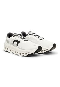 Zapatillas deportivas blancas con un upper de malla texturizada, acentos negros, un logo redondo y una suela acolchada distintiva que presenta segmentos huecos para la absorción de impactos.