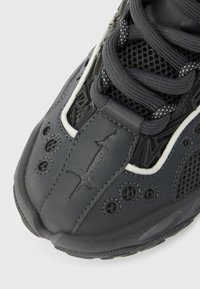 Μαύρο αθλητικό παπούτσι με πλέγματα, μαύρα κορδόνια με λευκές τελείες και ανάγλυφο λογότυπο σπαθί στην περιοχή των δακτύλων.