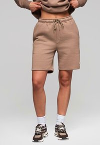 Shorts de sport beiges en tissu doux, dotés d'une taille élastique avec cordon de serrage et de coutures latérales. Portés avec des baskets volumineuses.