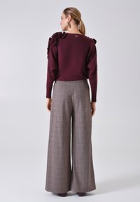 Burgunderfarbener Pullover mit Rüschendetail an der Schulter, kombiniert mit hochtaillierten, weit geschnittenen Karo-Hosen in Braun- und Beigetönen.