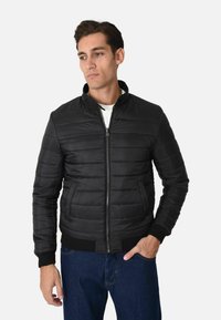 Giacca a vento nera con design trapuntato orizzontale, colletto alto, chiusura con zip e polsini a costine. Abbinata a jeans in denim blu.