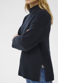 Femme portant un pull en maille côtelée navy avec un col haut, des manches longues, des fentes sur les côtés, et un jean bleu, se tenant les bras croisés.
