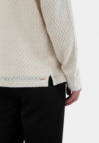 Texturerad krämfärgad tröja med ett diamantstickat mönster, med sidoflikar i fållen och en liten orange etikett på nedre vänstra sidan.