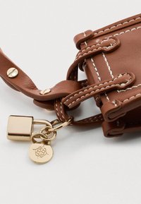 Braune Lederhandtasche mit weißer Näht, ausgestattet mit goldenen Hardware und einem Schloss. Beinhaltet einen Charme mit einer floralen Gravur, der durch eine Schlaufe befestigt ist.