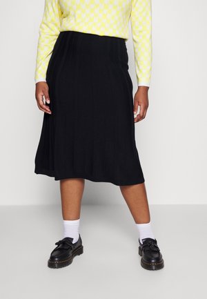 Falda de punto negra con canalé y dobladillo acampanado, combinada con un suéter amarillo con estampado y zapatos negros de suela gruesa y detalle de borlas.