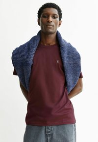 Bordeauxrode T-shirt met een klein logo, gecombineerd met een blauwe gestructureerde sjaal over de schouders gedrapeerd. Lichtgrijze spijkerbroek maakt de look compleet.